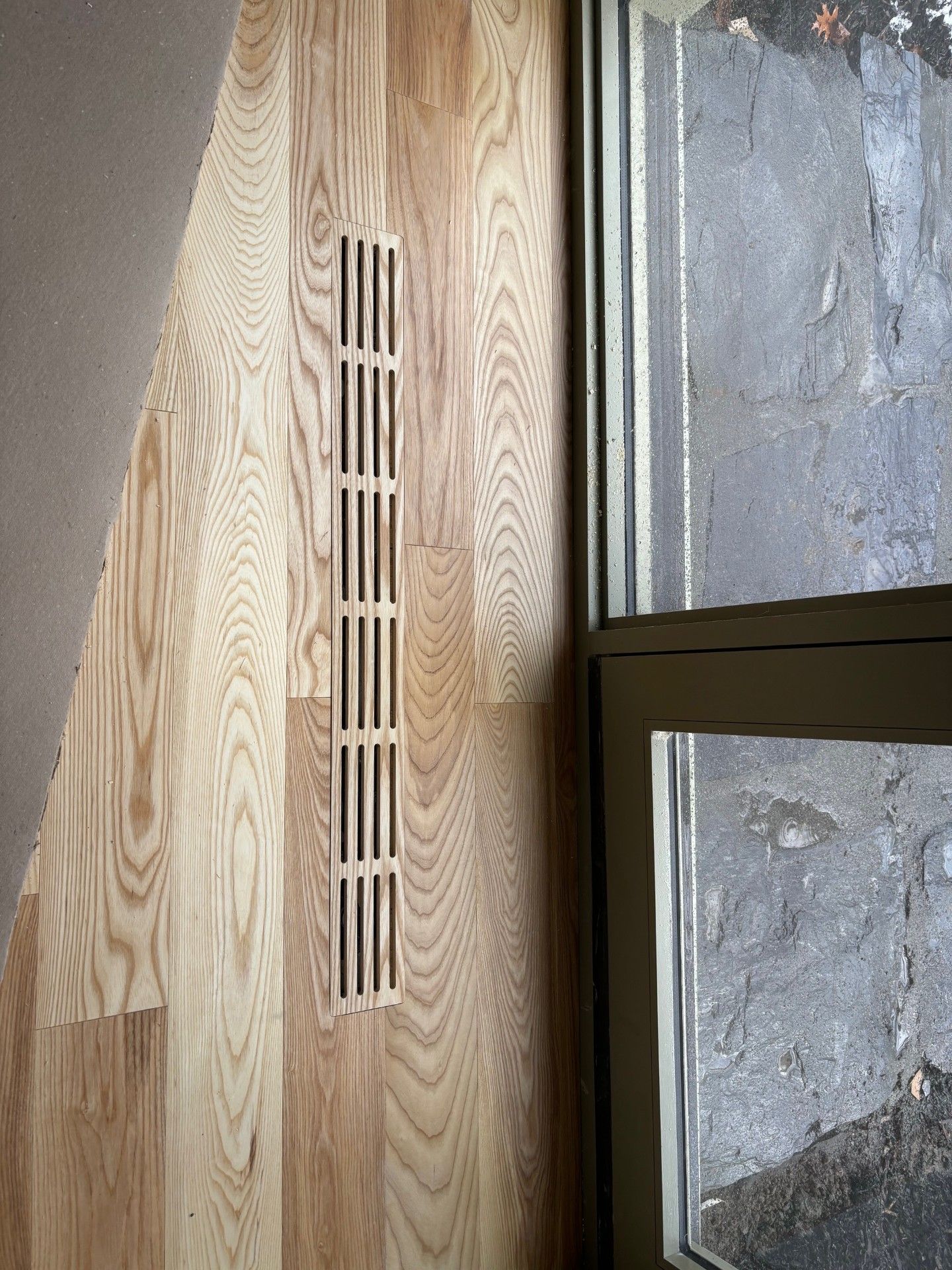 Plancher en bois avec une grille d'aération rectangulaire à côté d'une fenêtre.