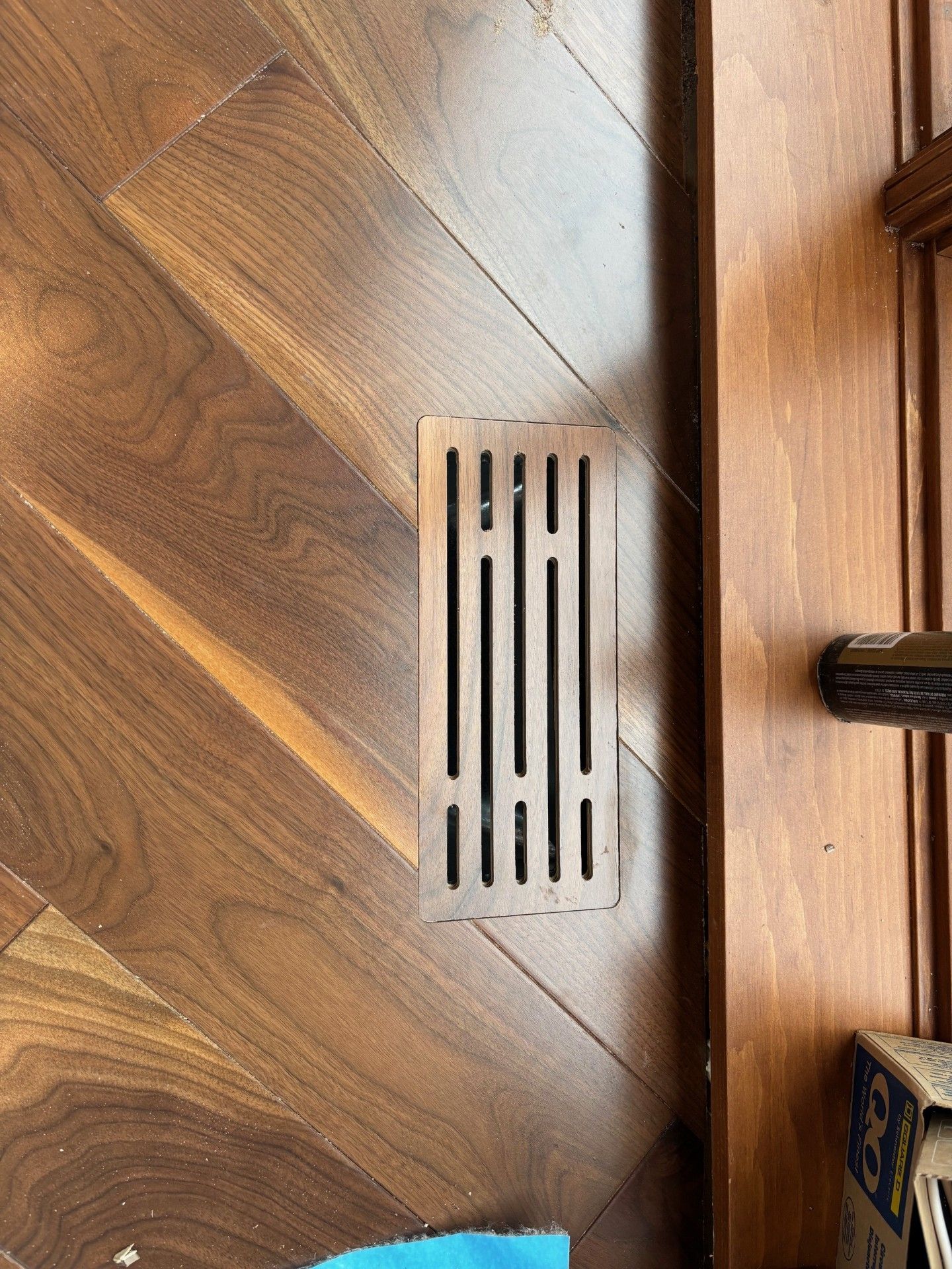 Grille d'aération en bois dans une pièce lambrissée. La grille est rectangulaire et présente une structure à fentes verticales.