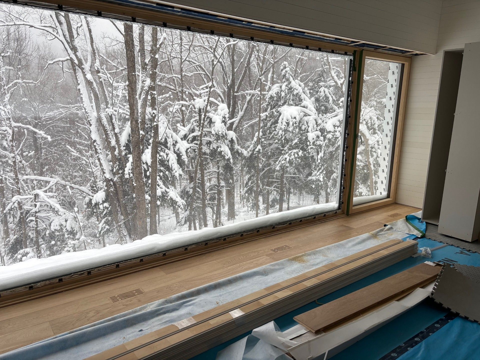 Grande fenêtre donnant sur une forêt enneigée. Boiseries et appui de fenêtre en bois clair, revêtement de sol intérieur assorti.