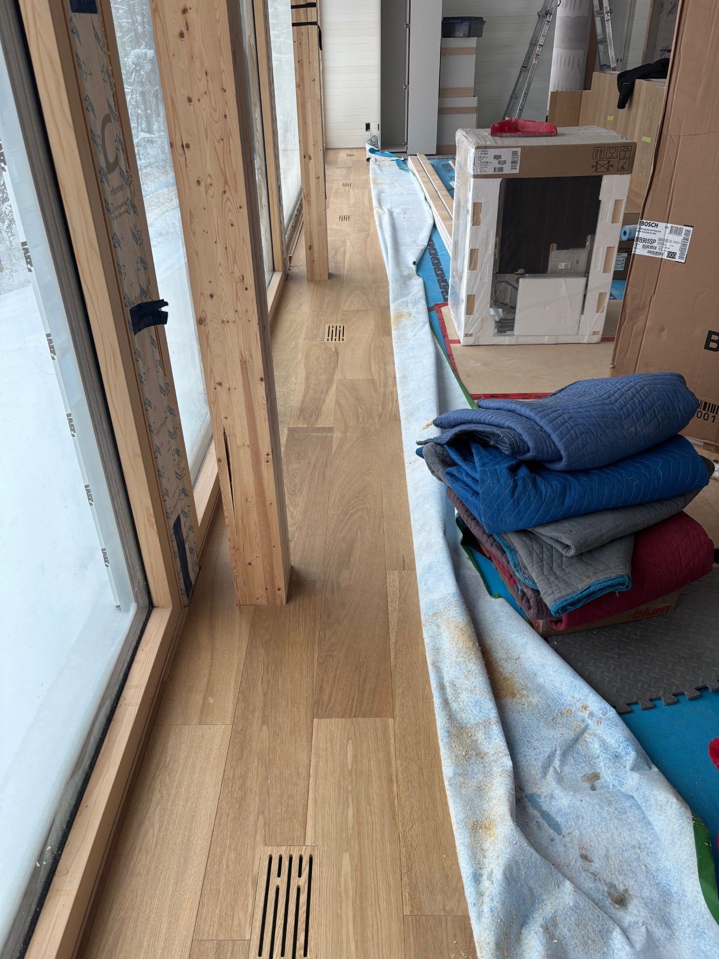 Couloir avec parquet, grandes fenêtres, pile de couvertures et cheminée partiellement construite.