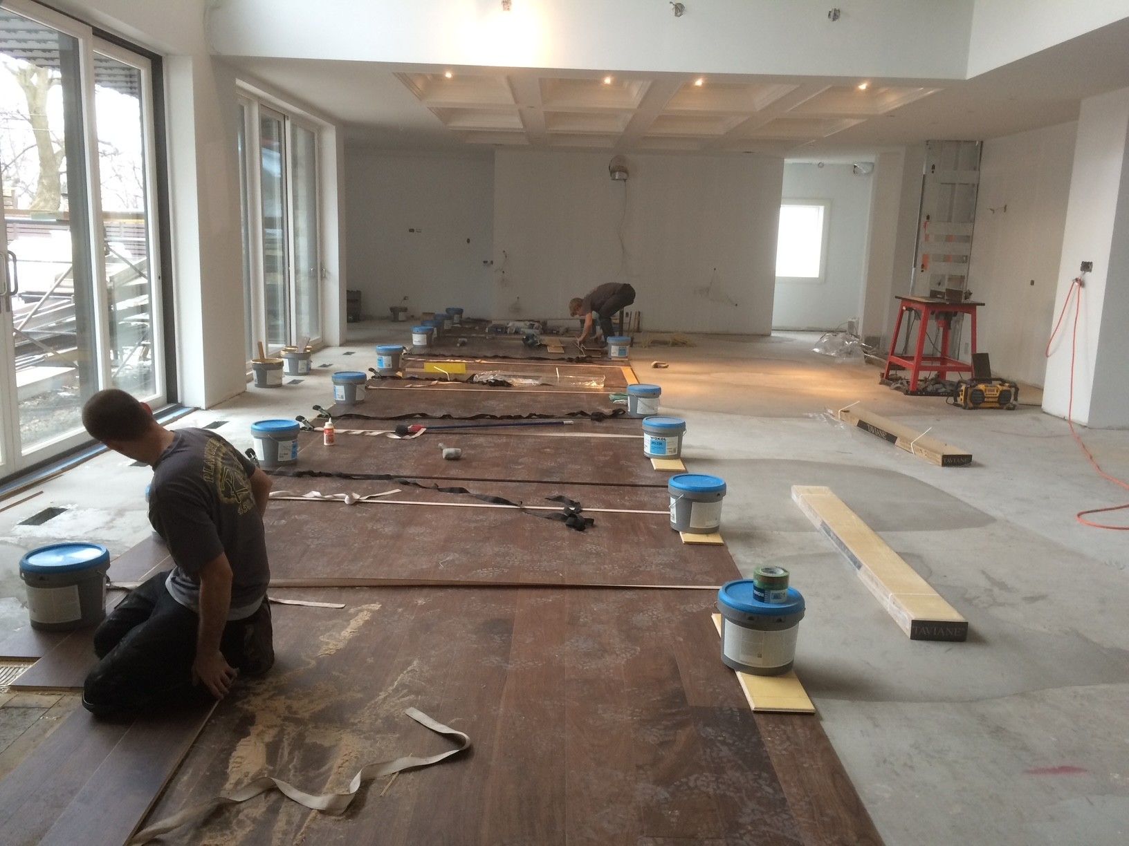 Deux ouvriers posent du parquet dans une grande pièce lumineuse. Des matériaux et des outils de construction sont présents.