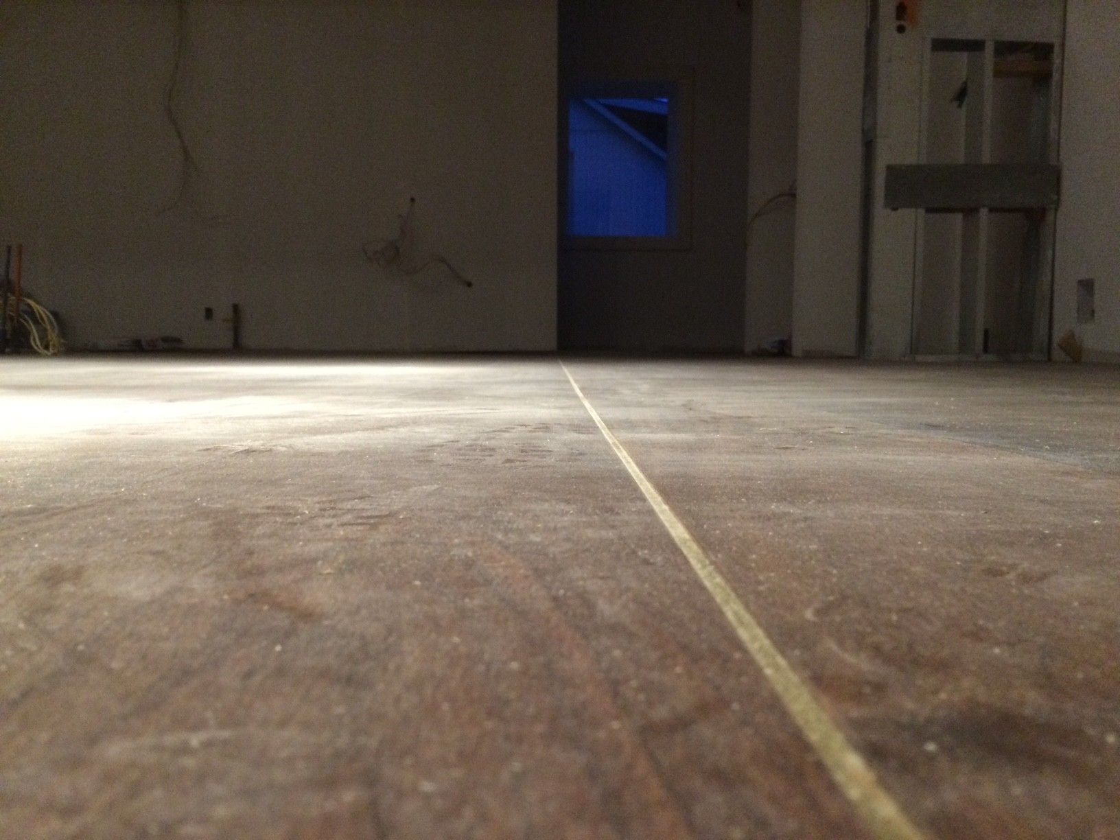 Vue en contre-plongée d'un parquet avec une ligne jaune, donnant sur une pièce faiblement éclairée avec une fenêtre bleue.