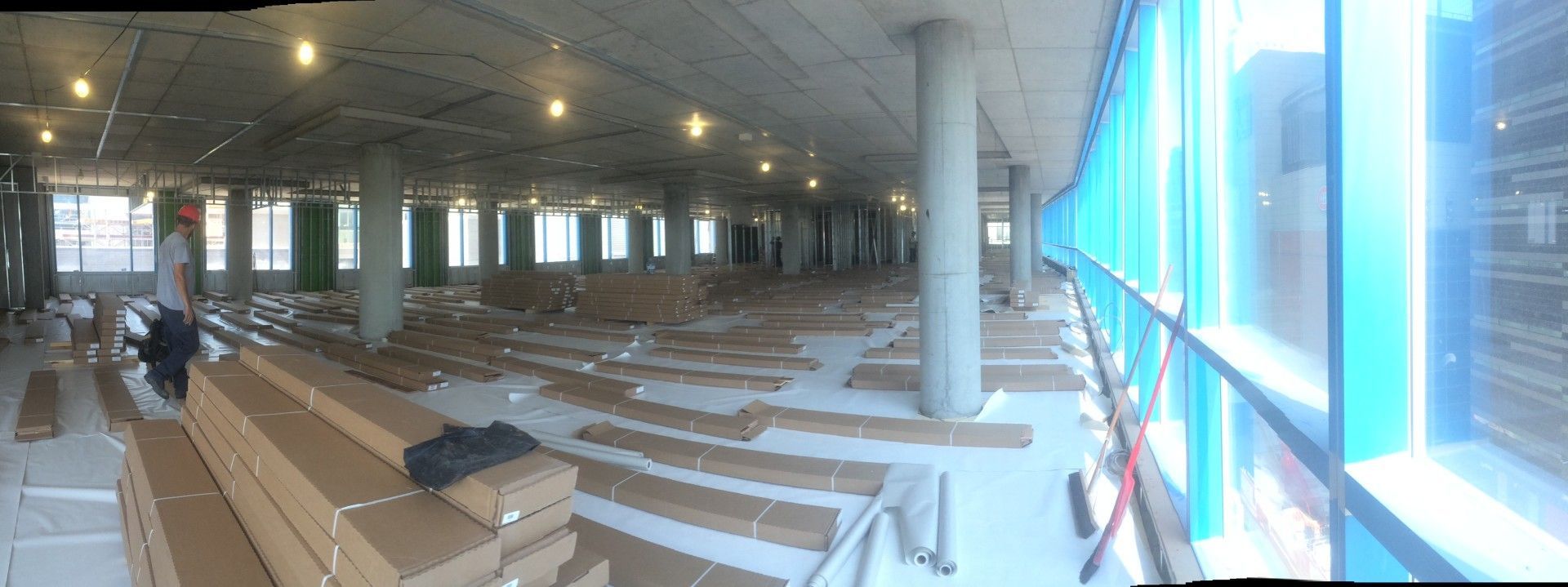 Intérieur d'un chantier de construction avec des caisses de matériaux de revêtement de sol, un ouvrier et des fenêtres incurvées teintées de bleu.