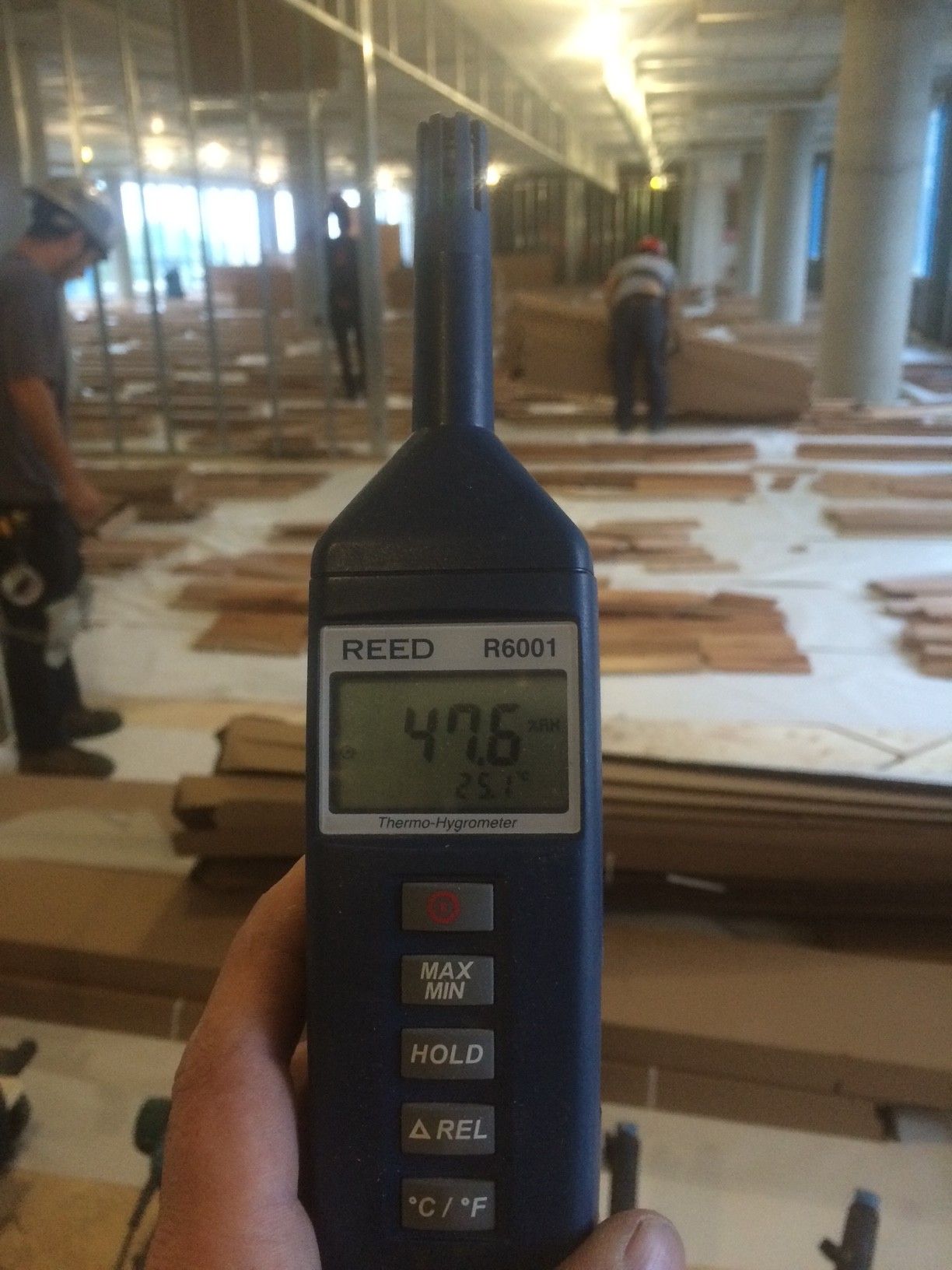 Main tenant un humidimètre sur un chantier de construction avec un plancher en bois, ouvriers présents. L'appareil indique 47,6.