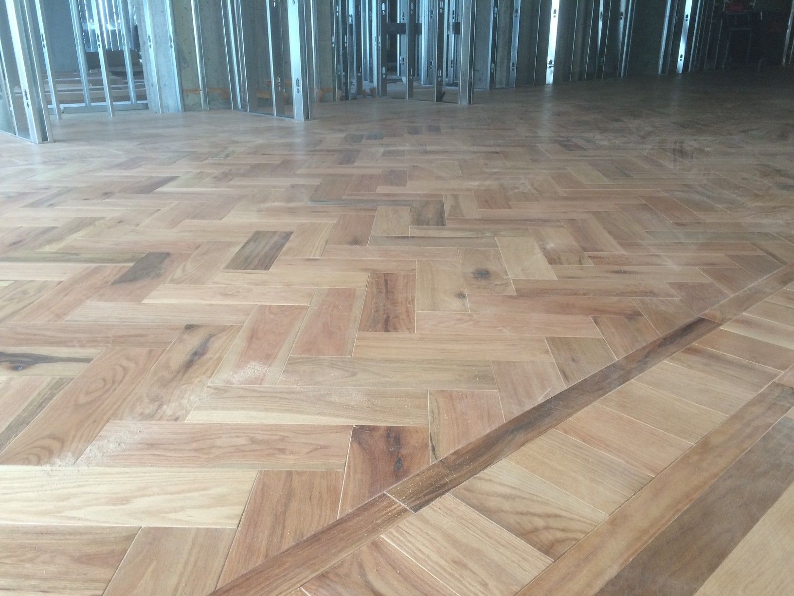 Parquet à chevrons, brun clair avec des plinthes foncées, reflétant la lumière d'une fenêtre à barreaux verticaux.