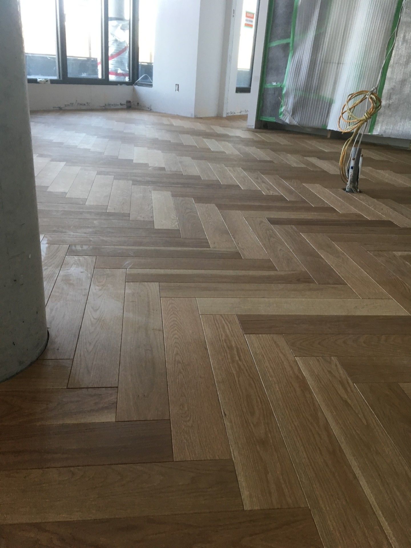 Parquet à chevrons dans une pièce en construction, avec un pilier en béton et une fenêtre visibles.