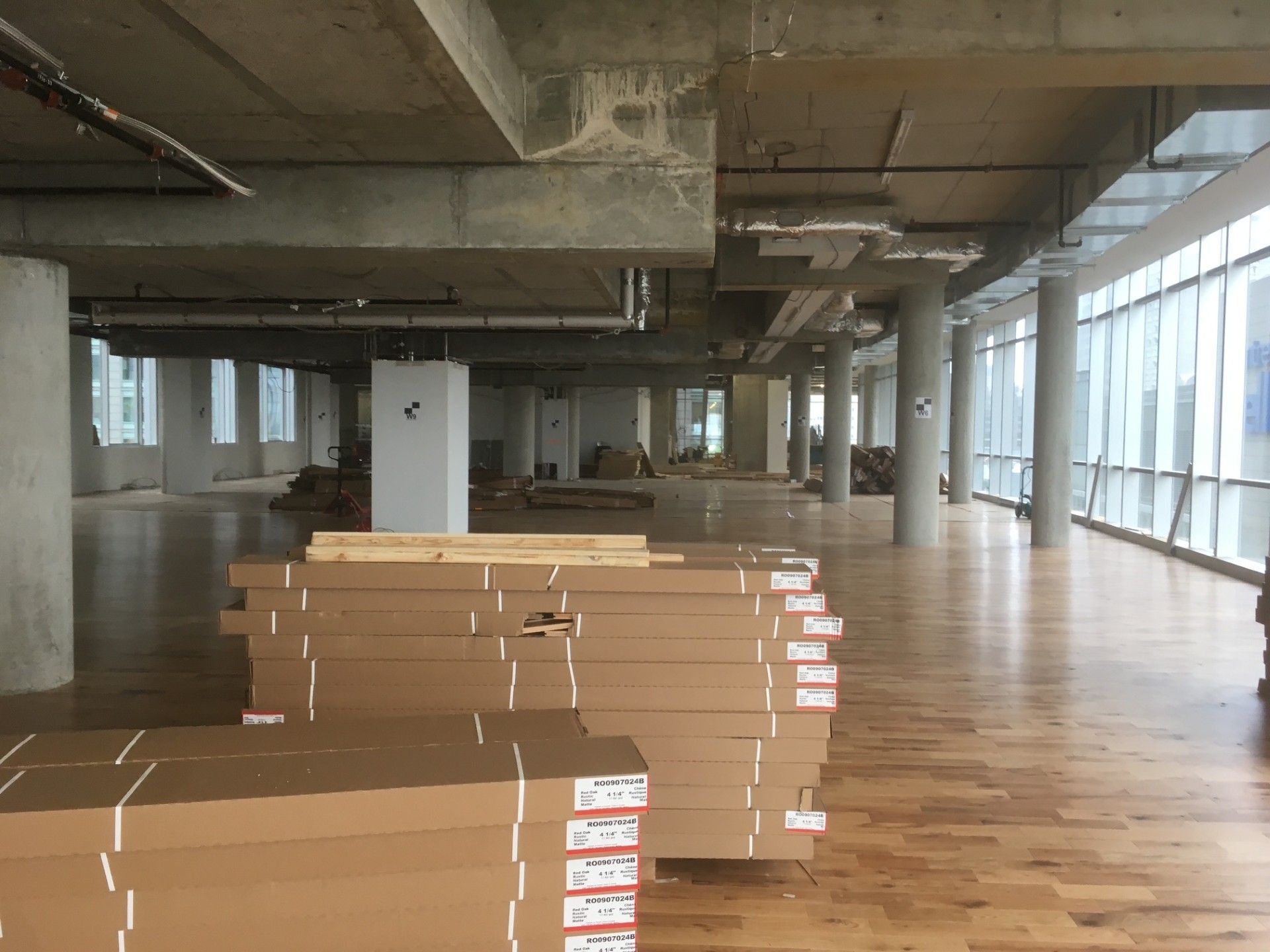 Intérieur d'un bâtiment inachevé avec des piles de cartons, un plancher en bois et de grandes fenêtres.