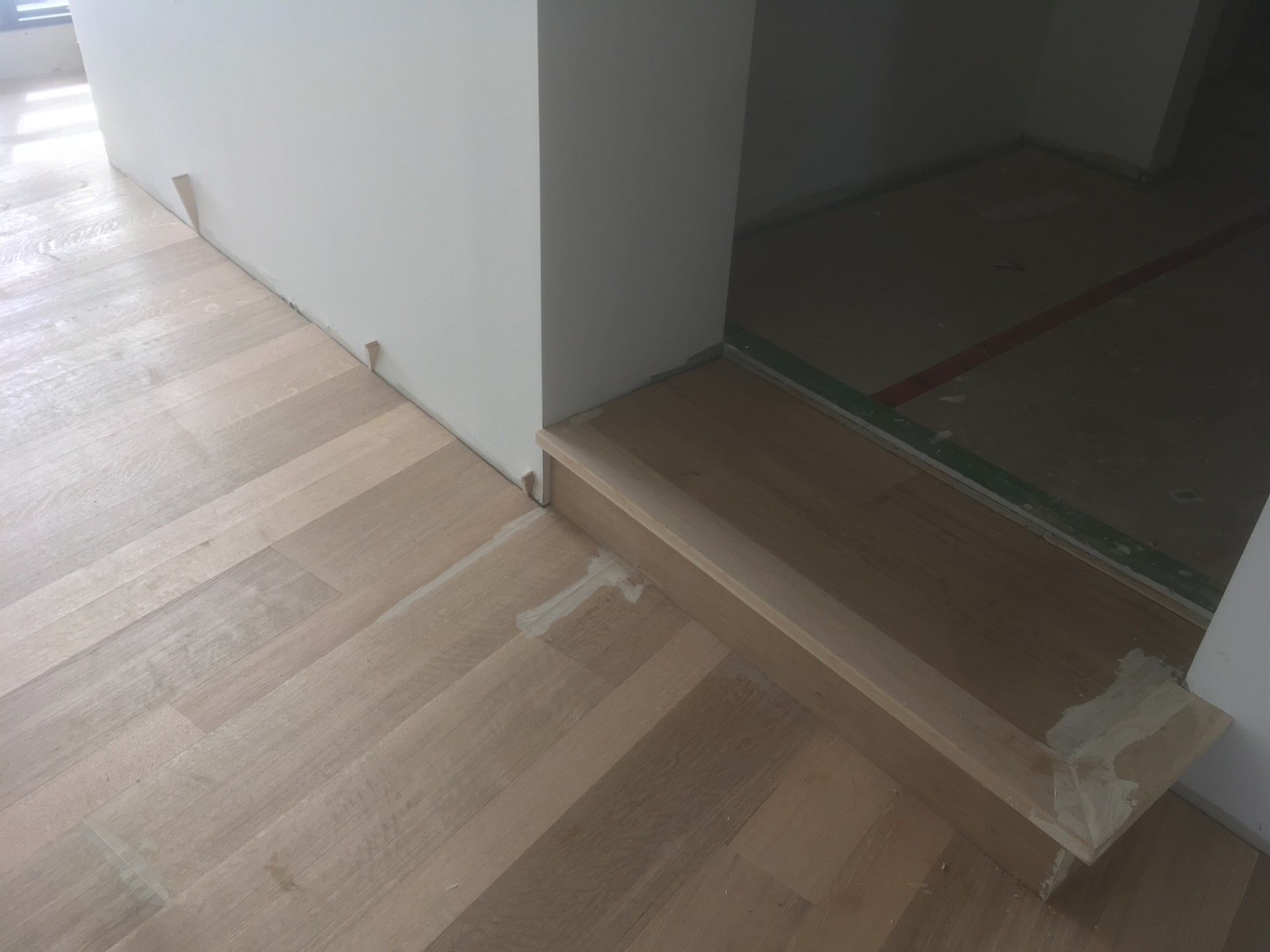 Plancher en bois franc avec une marche pour accéder à une porte. Bois clair, murs blancs et bâches de chantier visibles.