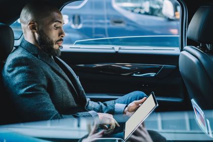 Man using a tablet in the back seat of a car, with a city street visible through the window.