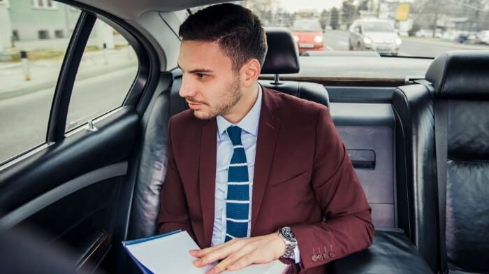 Man in a burgundy suit sitting in the back seat of a car, looking out the window and holding papers.