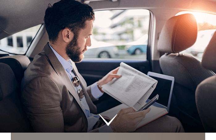 Man in a suit reading papers in the back seat of a car with a laptop beside him