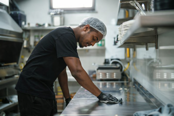 Kitchen worker in hairnet and gloves cleans stainless steel counter in bright, organized kitchen.