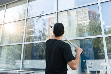 Man in black shirt and cap cleans large glass window with squeegee on sunny day; reflections of trees and building visible.