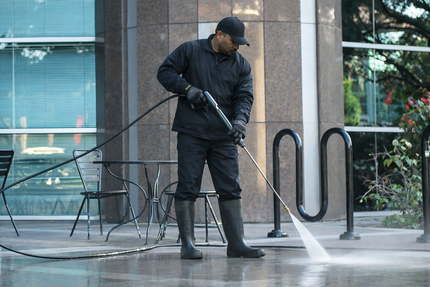 Man in black work attire power washes sidewalk in front of modern building, professional and focused.