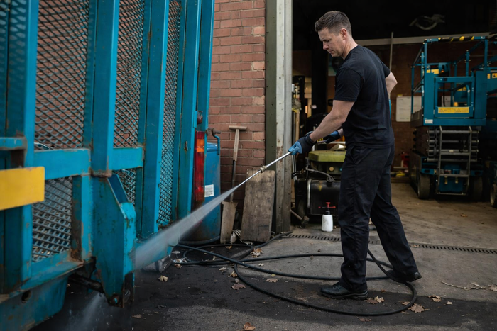 Man in dark clothing and gloves pressure washes blue metal gate outside gritty industrial building.