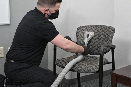 Person in black clothing and mask vacuums patterned office chair in focused, professional office setting.