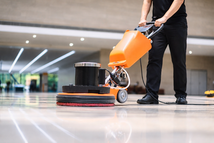 Person in black pants operates orange industrial floor buffer on shiny, polished floor in large, well-lit space.