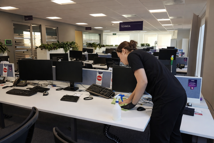 Person in black uniform cleans tidy, well-lit office with white desks and computers.