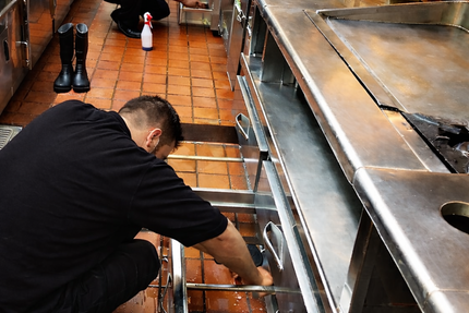 Two people in black outfits clean commercial kitchen one scrubs drawer, other cleans cabinet, showing focus and teamwork.