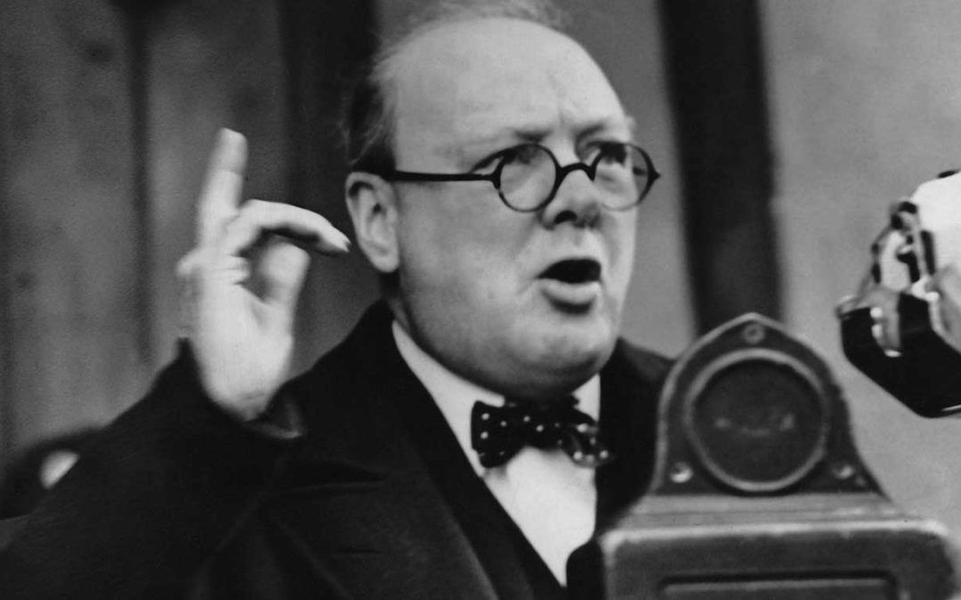 A man wearing glasses and a bow tie is giving a speech in front of a microphone.