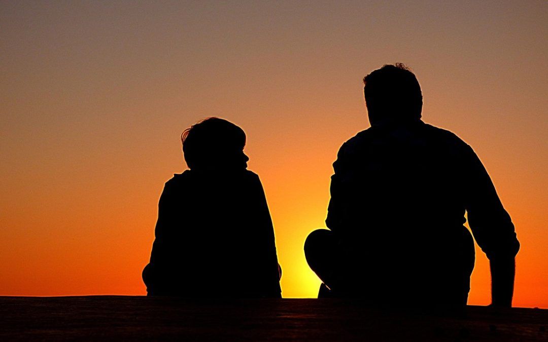 A man and a child are sitting in front of a sunset.