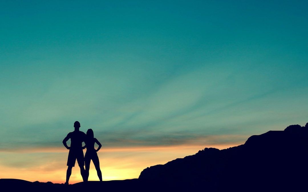 A man and a woman are standing next to each other on top of a hill at sunset.