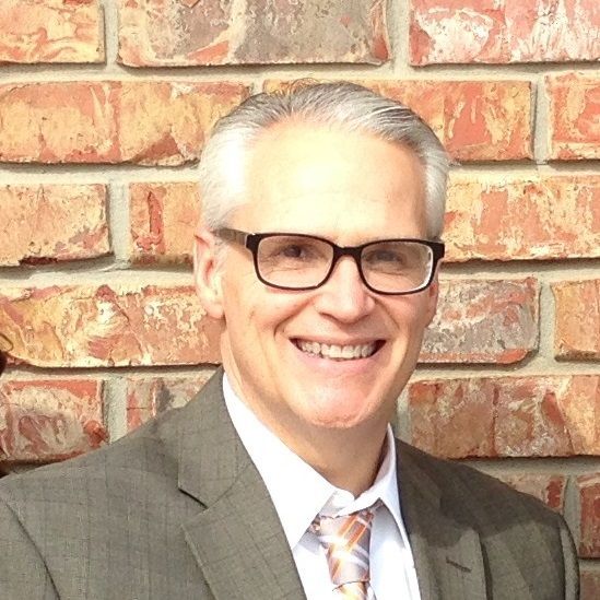 A man in a suit and tie is smiling in front of a brick wall.