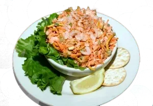 Shrimp salad in a white bowl on a plate, lettuce, crackers, and lemon.