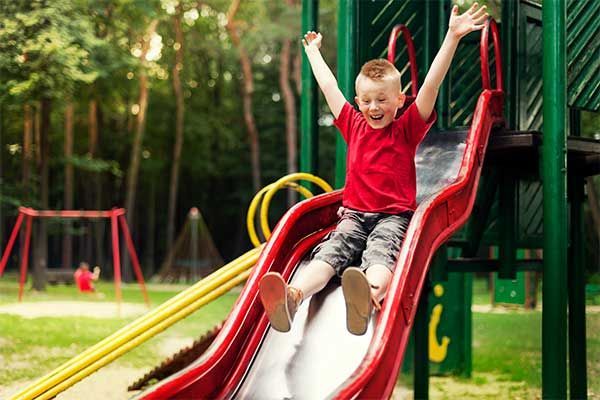 Child sliding down a red playground slide with arms raised in a green park
