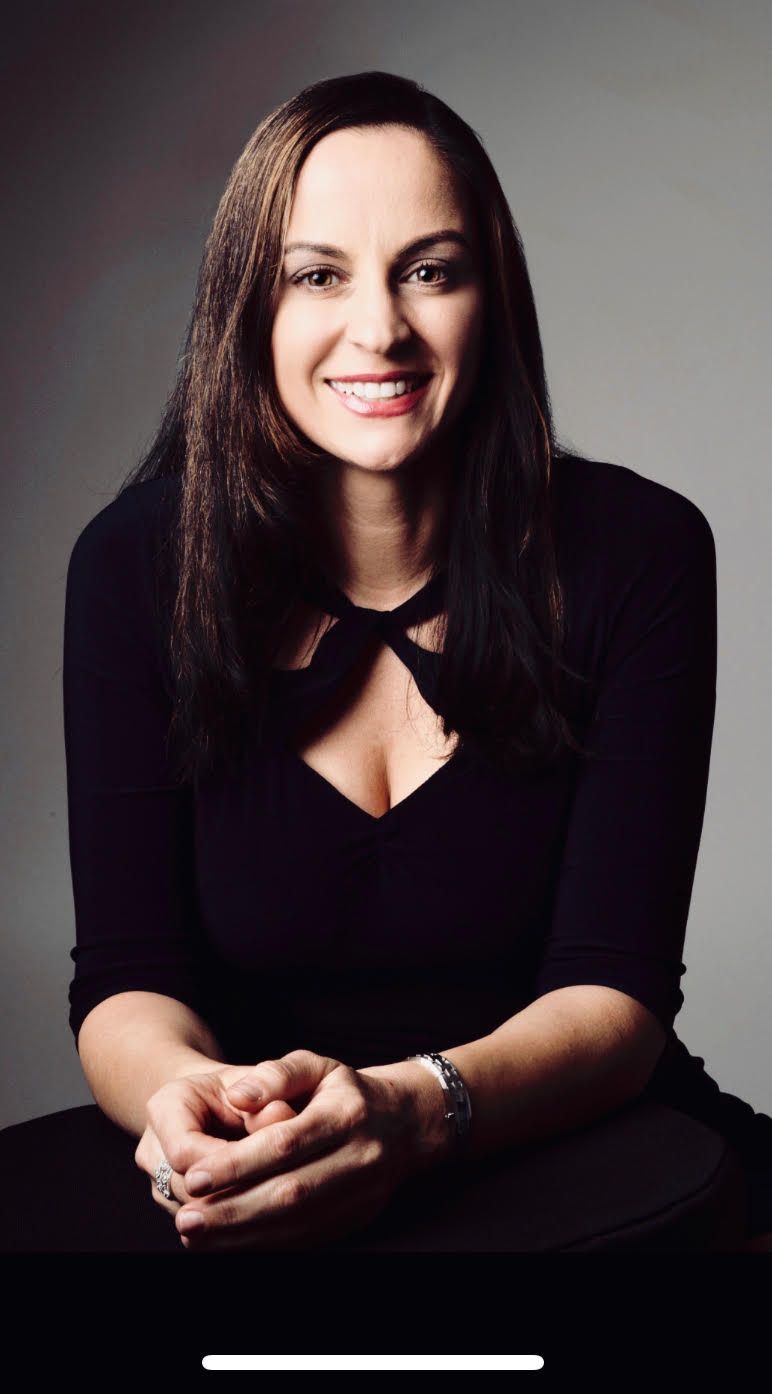 Portrait of Gordana smiling seated against a gray background, wearing a black top with a keyhole neckline.