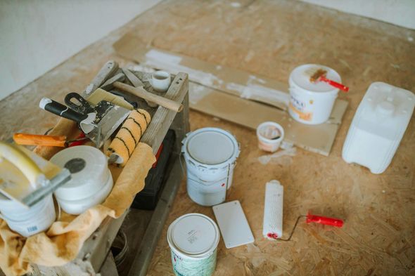 Painting tools on a wooden floor: paint cans, rollers, putty knives, drop cloth.