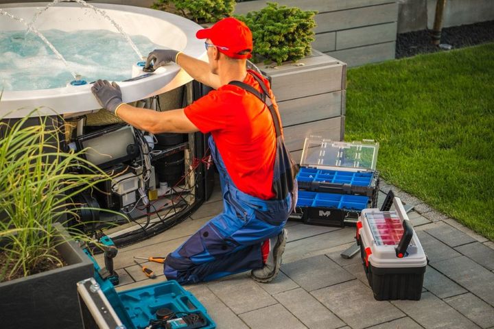 Technician in red cap and shirt repairs outdoor pool equipment beside a hot tub, with tools nearby