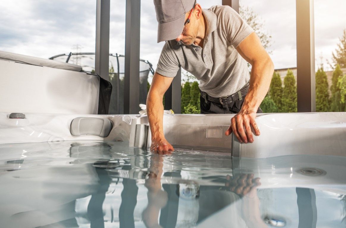 Man in a gray shirt cleaning water from a hot tub beside a house