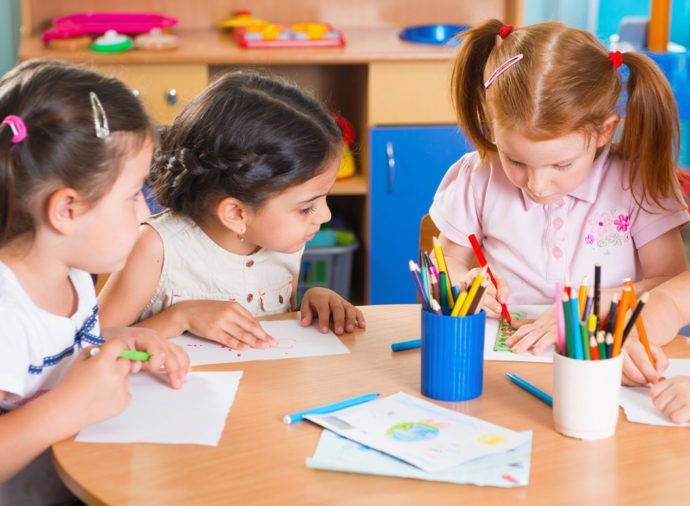 Three little girls are sitting at a table drawing with colored pencils.