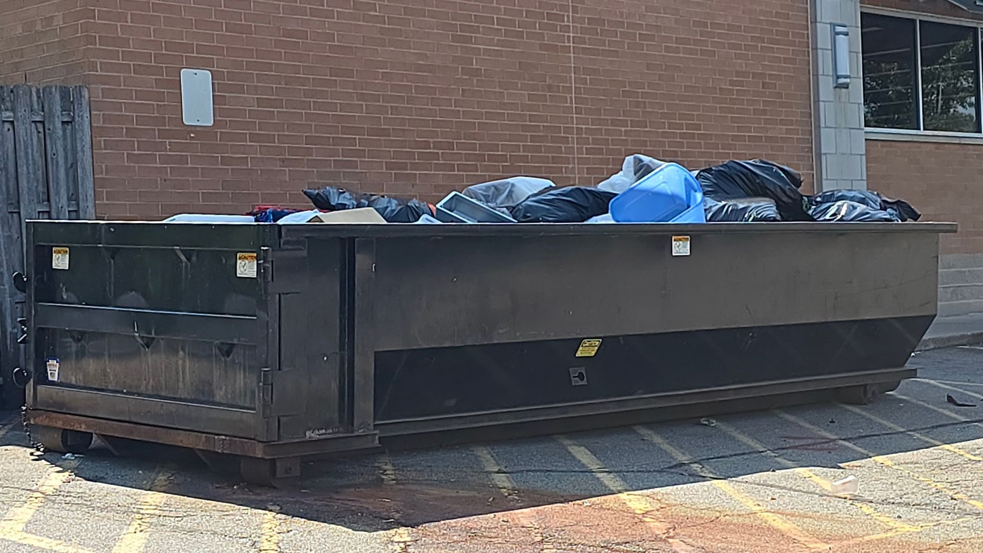 Black dumpster overflowing with trash bags, against a brick building.