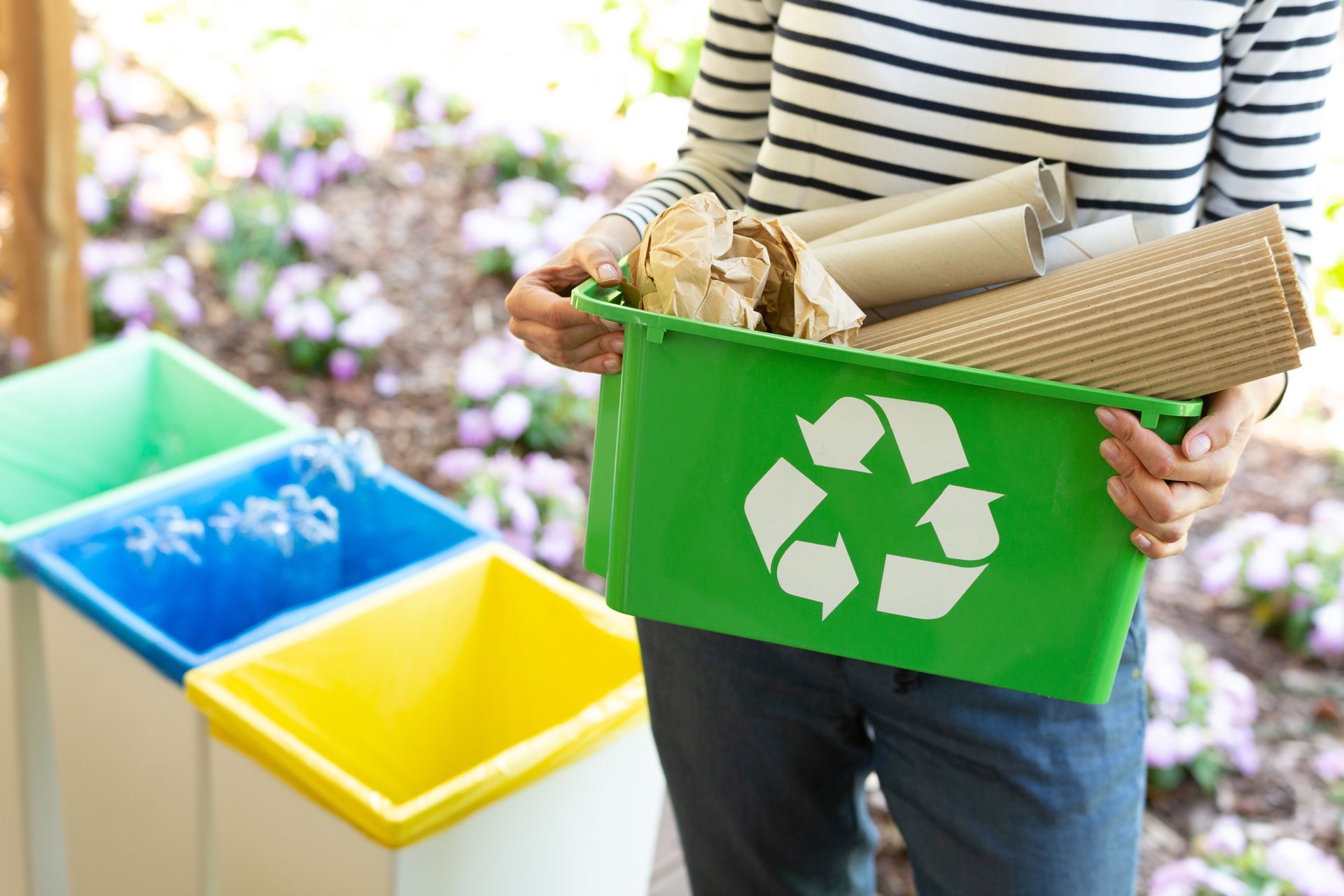 Person holding green recycling bin filled with paper and cardboard, near other colored bins.