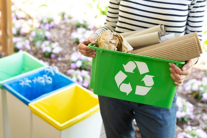 Person holding green recycling bin filled with paper and cardboard, near other colored bins. Person holding green recycling bin filled with paper and cardboard, near other colored bins.