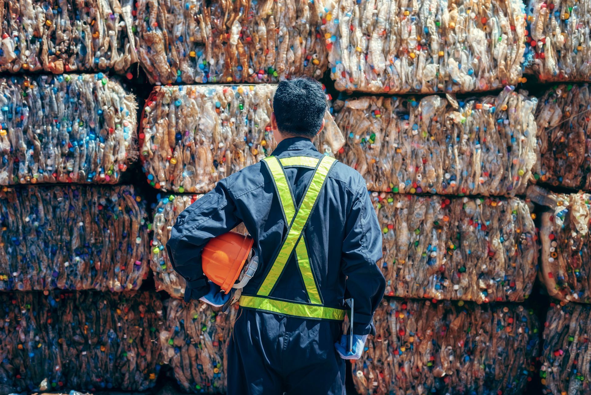 Man in safety vest holding a hard hat stands before stacked, compressed bales of recycled material.