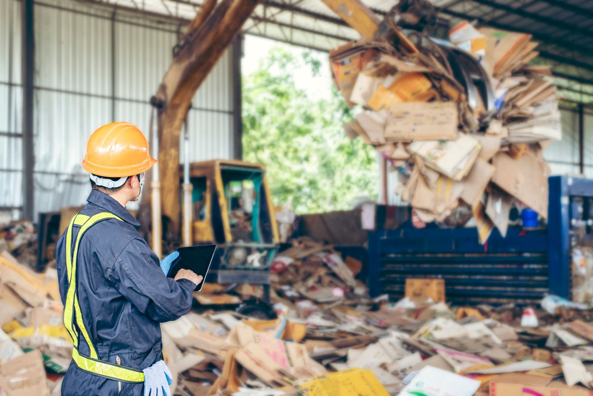Worker in safety gear inspects recycling pile with tablet, excavator in background.