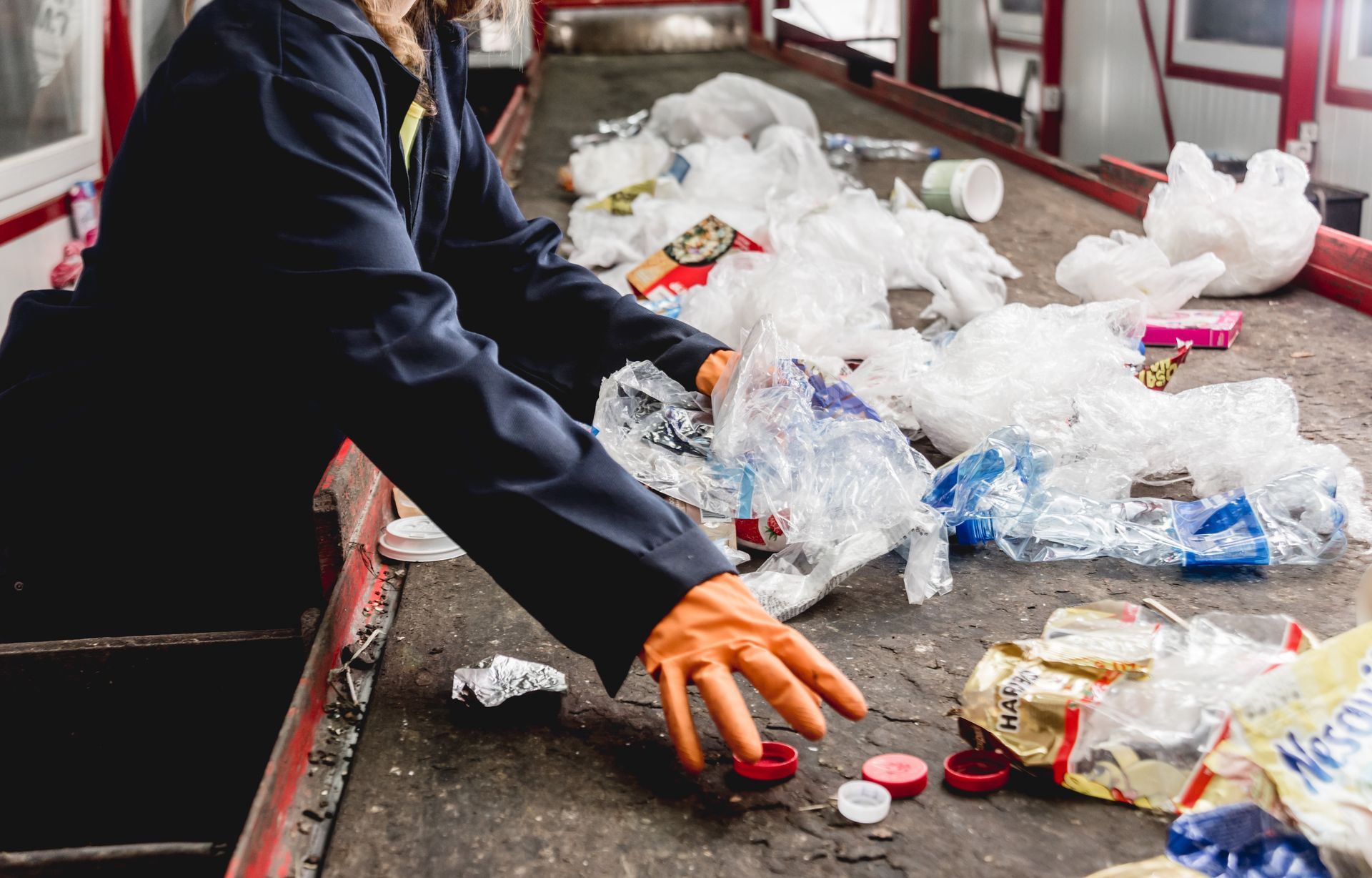 Person sorting plastic waste on a conveyor belt, wearing gloves.