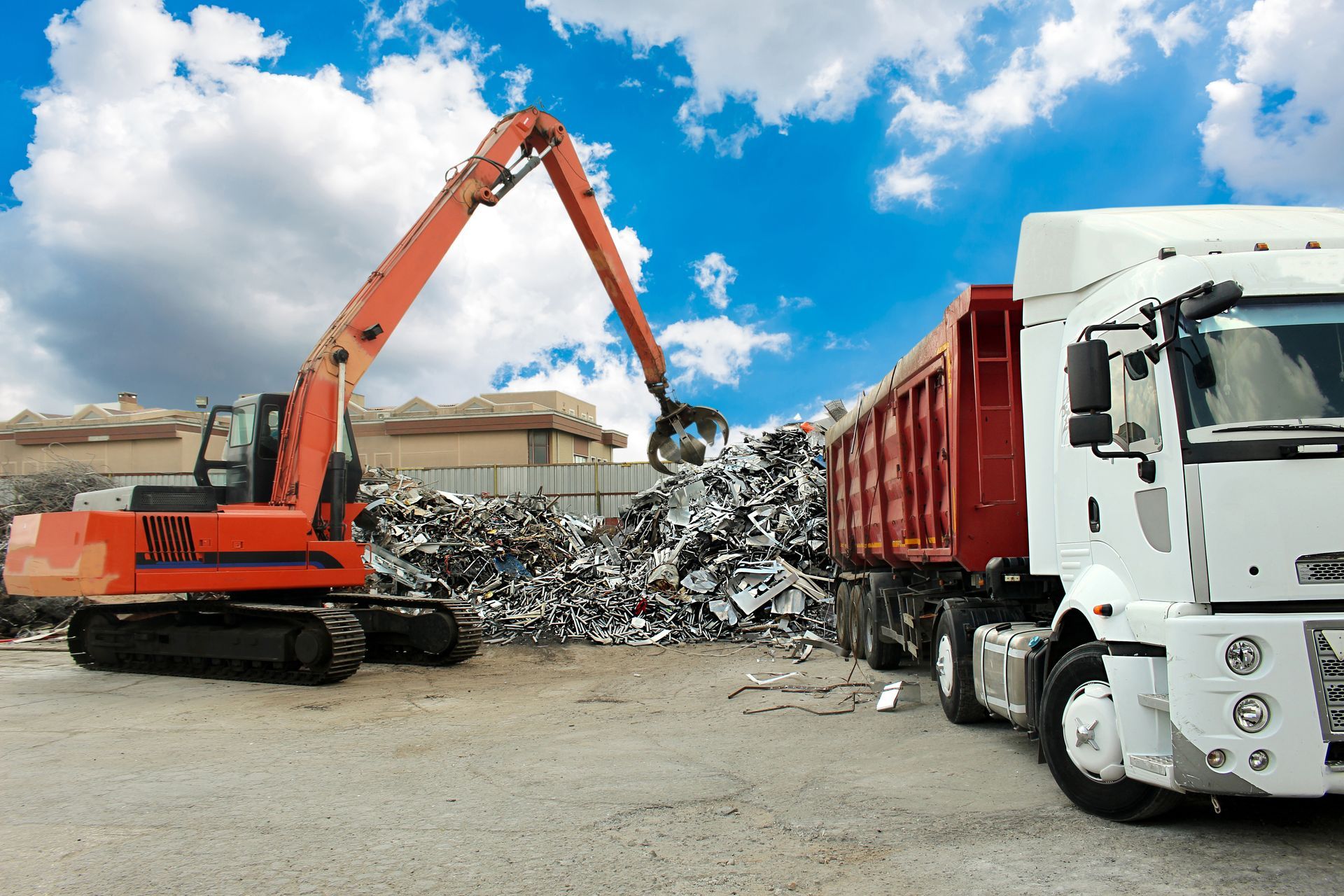 Orange excavator loading scrap metal into a red trailer truck at a recycling yard under a cloudy sky.