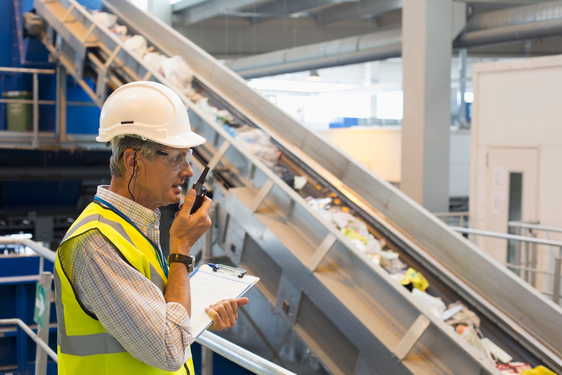 Person in safety gear using a radio, observing a conveyor belt in a waste facility.
