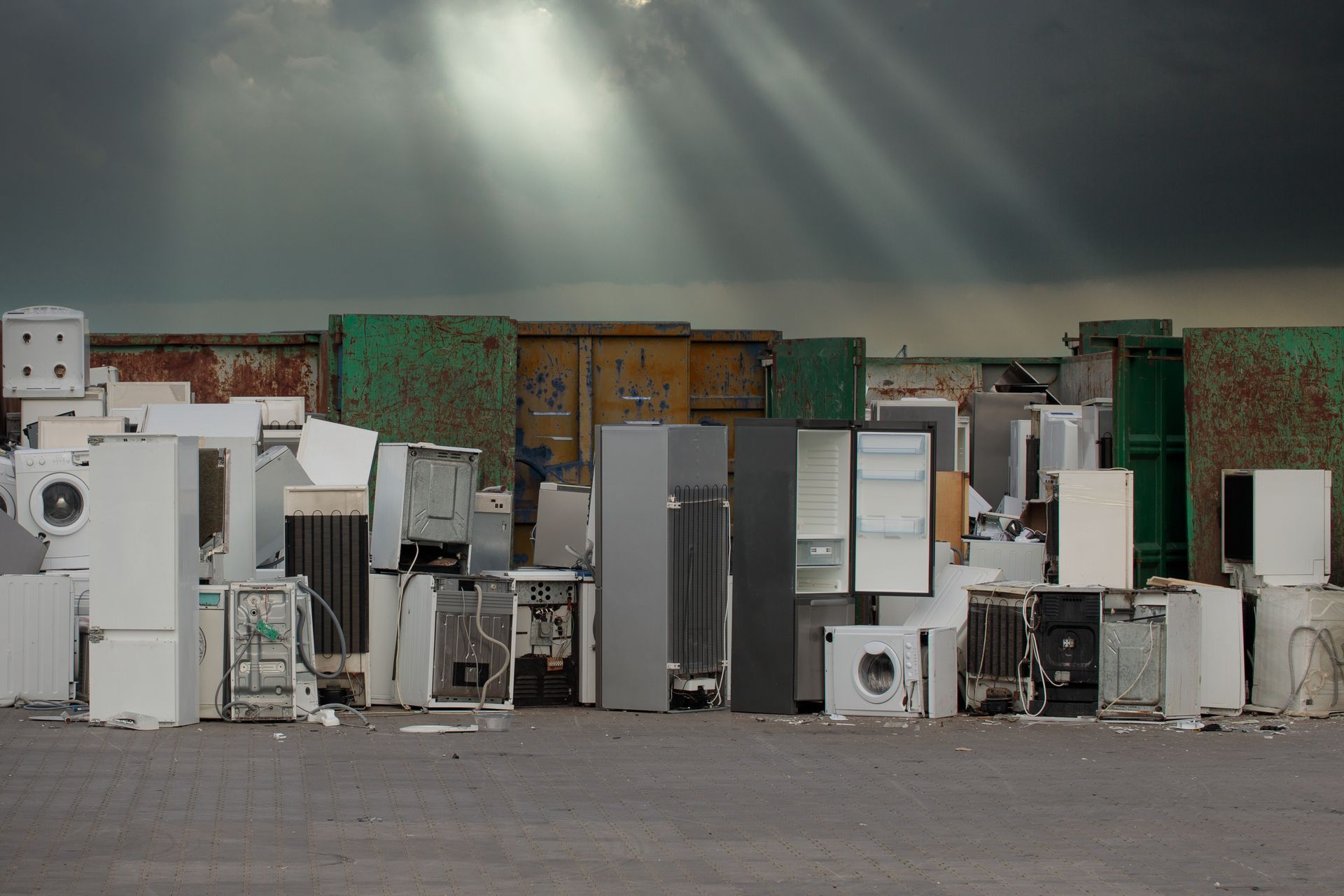 Old appliances in a junkyard under a dark sky with sun rays shining through.