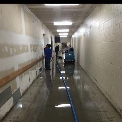 Hallway flooded with water, workers operate equipment. Walls are white, some damaged.