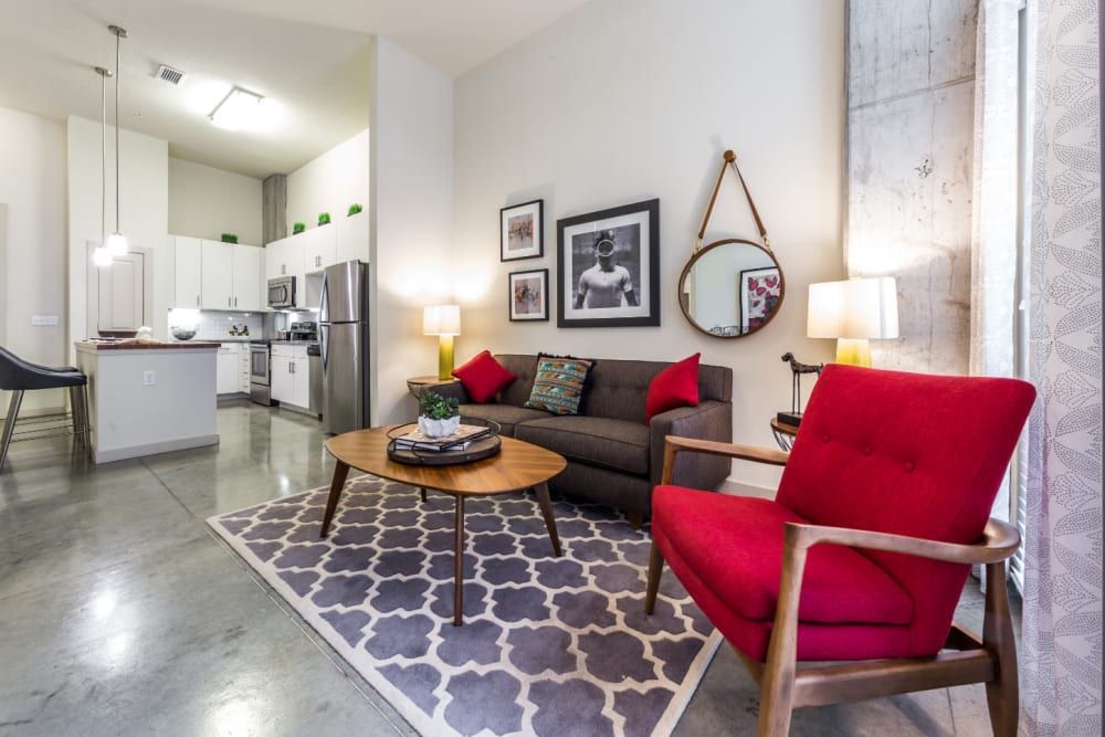 Modern living room interior with an open kitchen design. Features include sleek stainless steel appliances, white cabinetry, and a central island in the kitchen area. The living space is furnished with a dark brown sofa, two red accent chairs, a round wood coffee table, and a patterned gray and white area rug. Decorative elements include framed art on the walls, a round mirror with a wooden frame, and a variety of pillows. Concrete flooring runs throughout the space, and natural light comes from large windows dressed with sheer curtains at Marq on Burnet in Austin, TX.