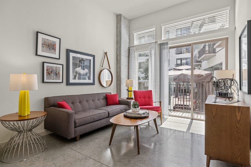 A modern living room featuring a gray sofa with red cushions, a single red armchair, and a round wooden coffee table flanked by metal lampstands with yellow shades. The room is decorated with framed art on the walls and a large oval mirror. Large windows open to a balcony overlooking an outdoor seating area. The space is illuminated with natural light, giving it a bright and airy feel at Marq on Burnet in Austin, TX.