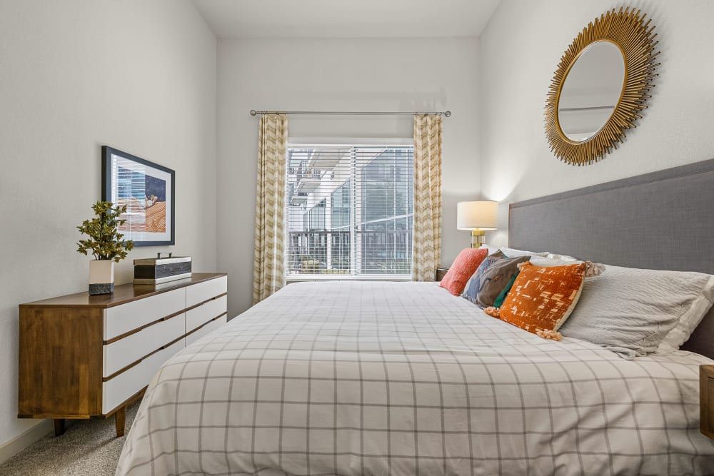 A modern, neatly arranged bedroom with a large window allowing natural light. The room includes a comfortable bed with a checkered duvet cover and several pillows in shades of grey, white, and burnt orange. A stylish, round, sunburst mirror hangs on the wall beside the bed, which is flanked by a wooden nightstand and dresser with white drawers featuring horizontal brown handles. Decorative elements include a potted plant and a framed picture on the dresser, patterned curtains, and a table lamp with a white shade. The overall color scheme is neutral with warm accents at Marq on Burnet in Austin, TX.