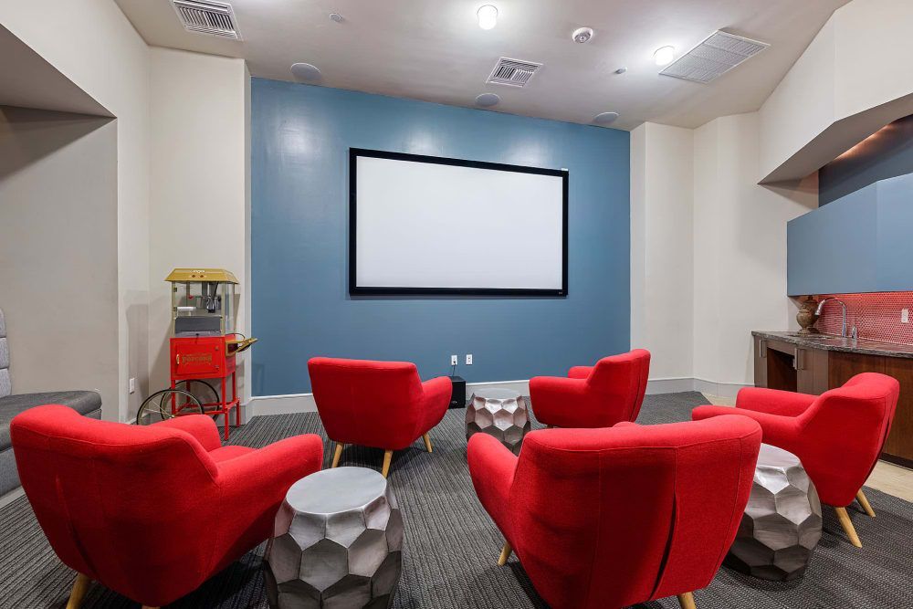 A modern home theater room with red armchairs arranged facing a large blank projector screen on a blue wall. To the left is a vintage-style red and yellow popcorn machine, and to the right is a kitchenette area with a sink and red mosaic tile backsplash. Overhead lighting and a grey carpet complete the cozy viewing space at Marq on Burnet in Austin, TX.