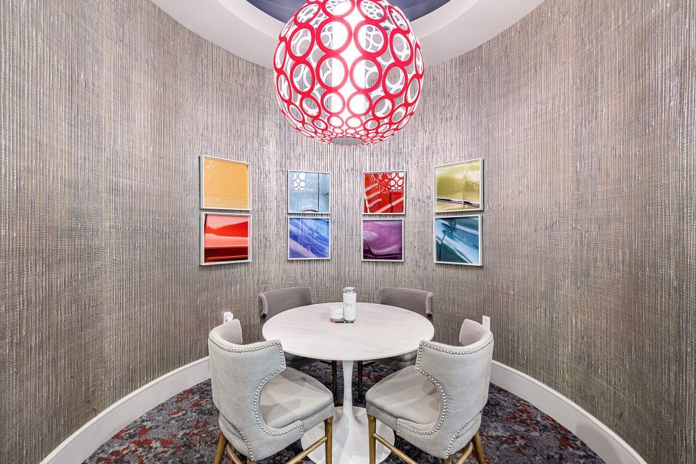 A modern lounge area featuring a round table with four grey upholstered chairs, a vibrantly patterned red and white spherical pendant light, and a collection of six colorful abstract art pieces displayed on a textured wall covering. The floor is adorned with a multicolored area rug at Marq on Burnet in Austin, TX.
