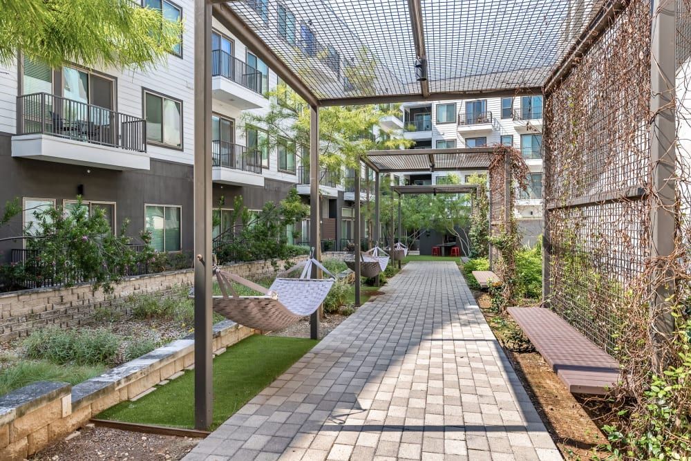 A modern apartment complex courtyard with landscaped gardens, stone walkways, and hammocks hanging under a metal trellis. The building consists of multi-story units with balconies overlooking the communal area. The courtyard is designed with seating benches along the path, lush plants, and a protective mesh overhead at Marq on Burnet in Austin, TX.